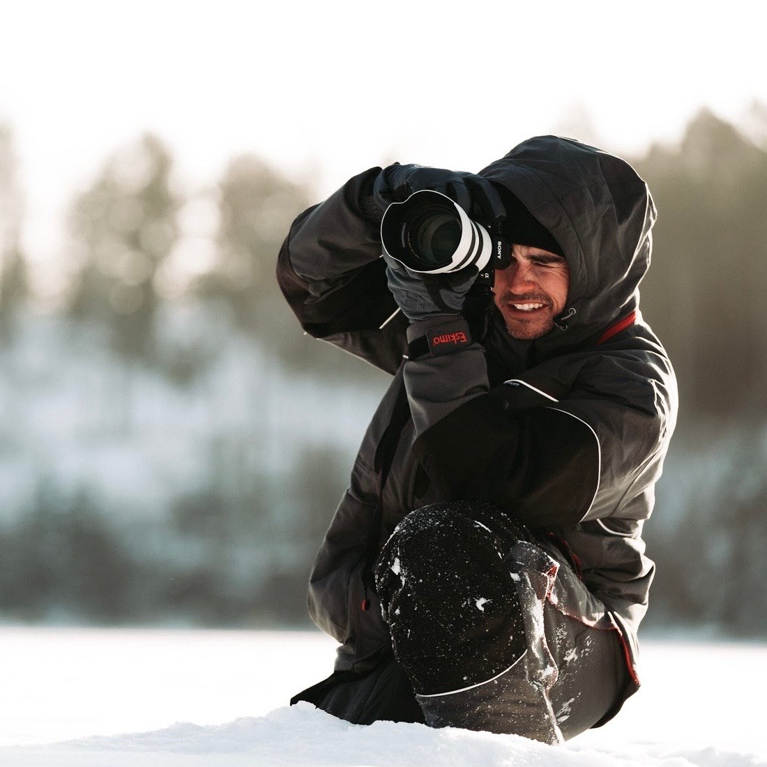 Ben Alcocer kneeling in deep snow in a heavy parka, camera raised to his eye, a treeline blurred behind him in flat winter light.