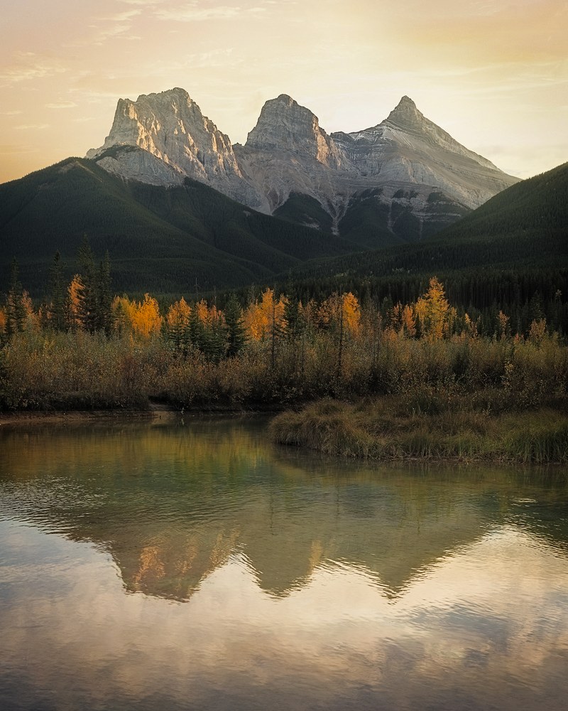 Three rock summits catch first light above a forested slope, autumn aspens and their reflection in the river below.