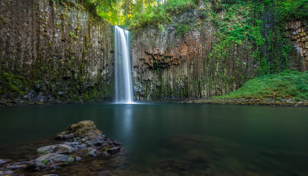 A single waterfall drops into a still dark pool inside an amphitheatre of columnar basalt, a low rock shelf in the foreground.