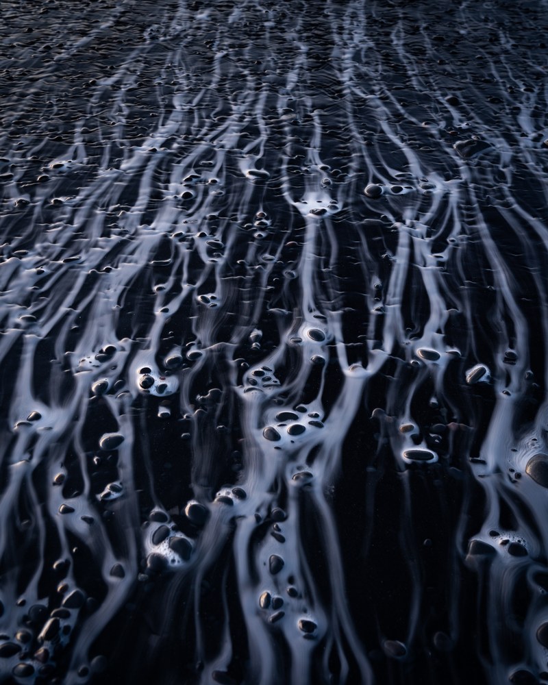 Looking down on black sand where retreating foam has drawn pale branching veins around scattered dark pebbles.
