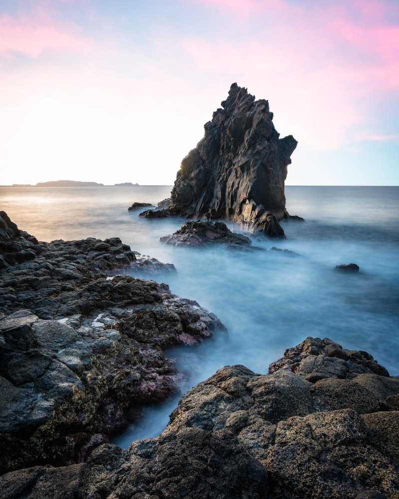 A jagged volcanic sea stack rises from surf blurred smooth by a long exposure, dark boulders in the foreground under a pale pink dusk sky.