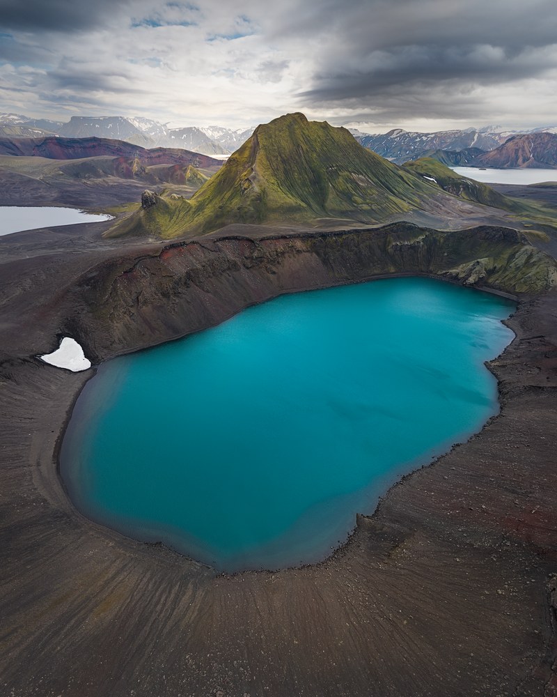 A turquoise crater lake sits inside a dark ash cone, a green-flanked peak and snow-streaked highlands beyond.