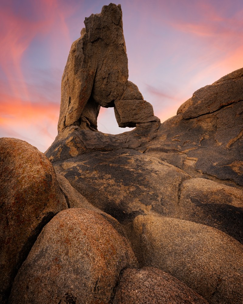 A blade of granite balances above a small arch in a pile of rounded boulders, pink cloud streaking the sky behind.