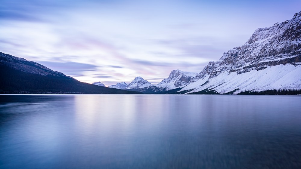 A still lake runs out to distant snow peaks, a long snow-covered ridge closing the right side under pale blue light.