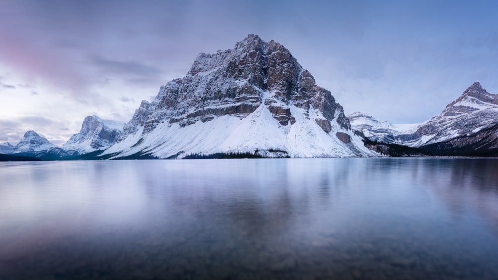 A broad snow-covered peak stands over a still lake that holds its reflection, smaller summits ranged on both sides.