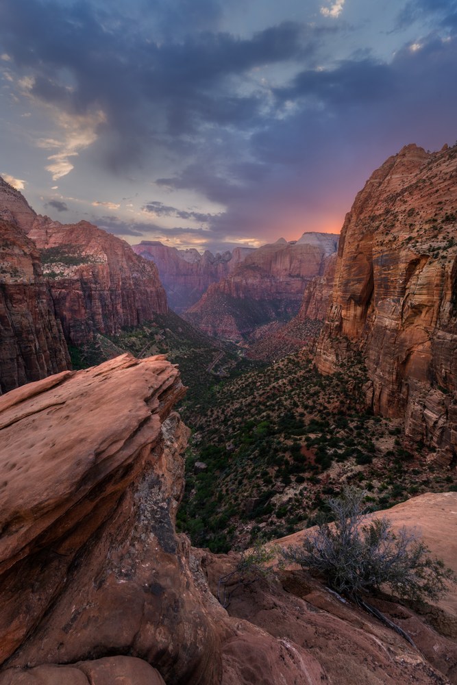 A bare rock ledge juts over a wide canyon of layered sandstone cliffs, storm cloud breaking into colour above the far rim.