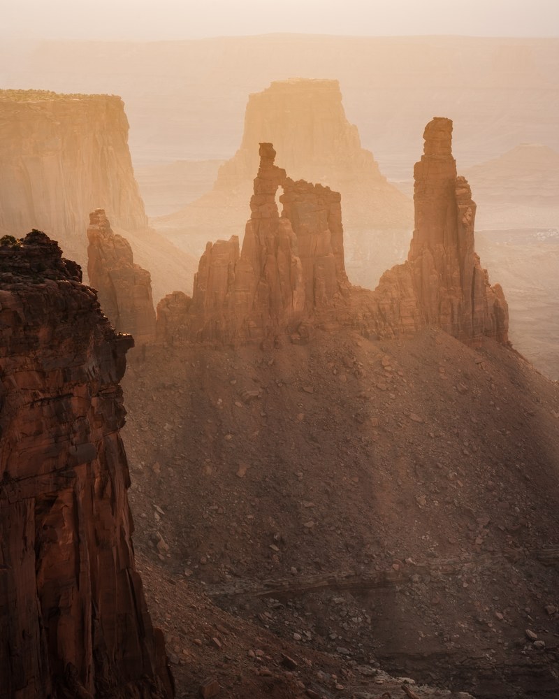 Slender rock spires stand on a rubble slope, ranks of mesas fading behind them in heavy golden haze.