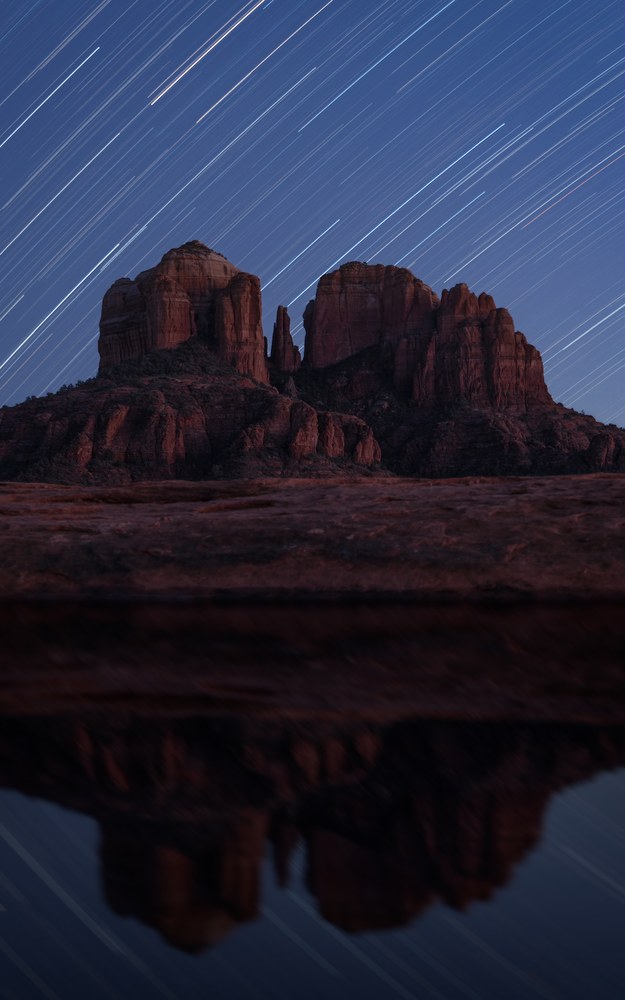 Star trails streak in parallel lines above a red butte, the whole scene repeated in still water in the foreground.