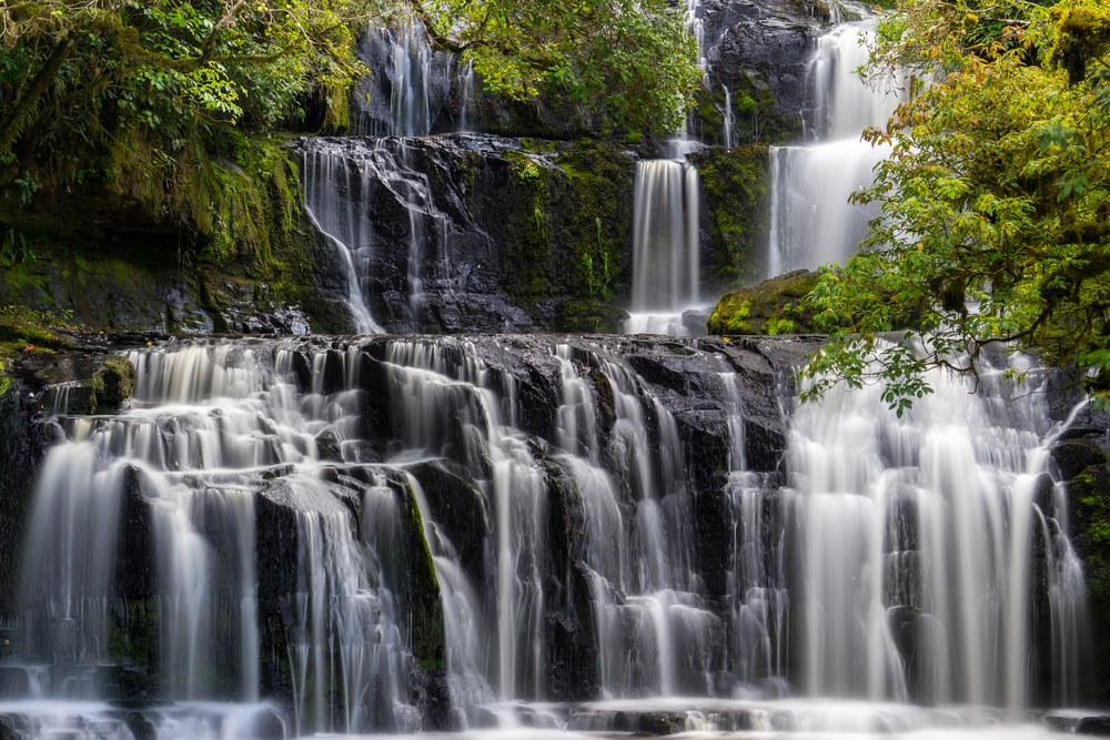 Water spreads in dozens of small threads down two wide tiers of dark rock ledges, framed by green bush.
