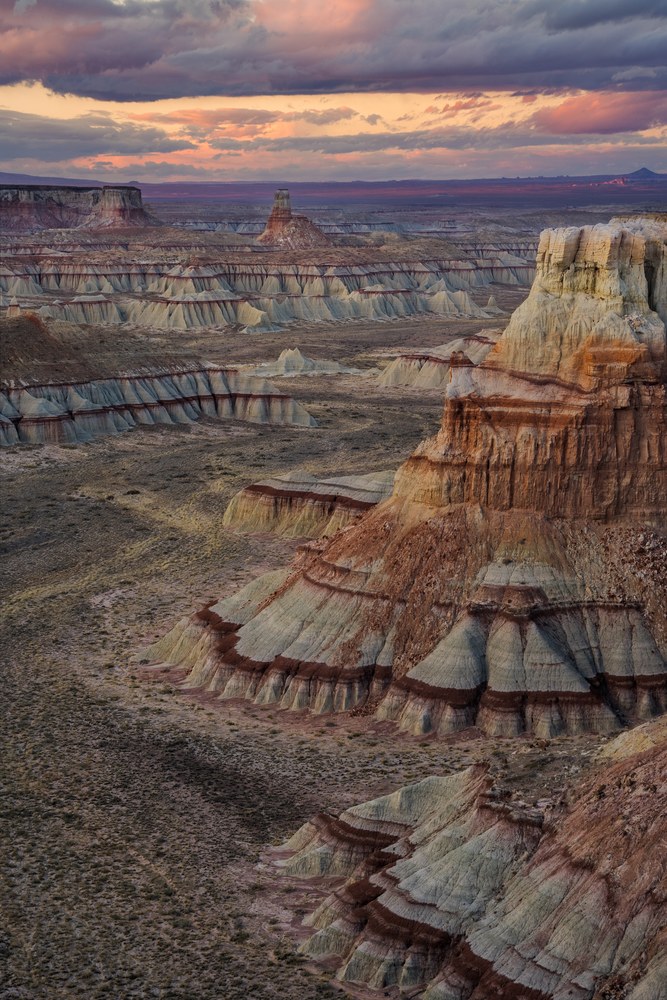Banded badland ridges and hoodoos step away toward a flat horizon under heavy cloud breaking into pink.
