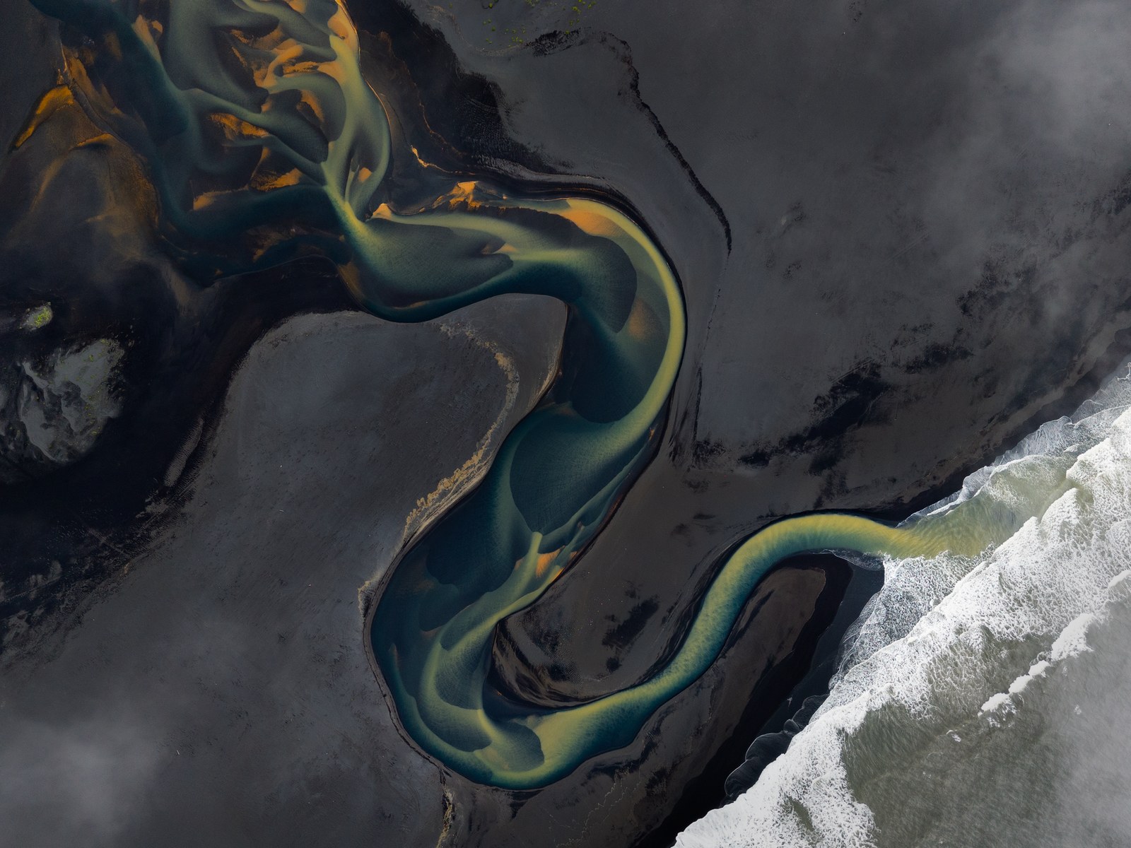 Seen from above, a glacial river braids green and gold across black sand before meeting a white line of surf.