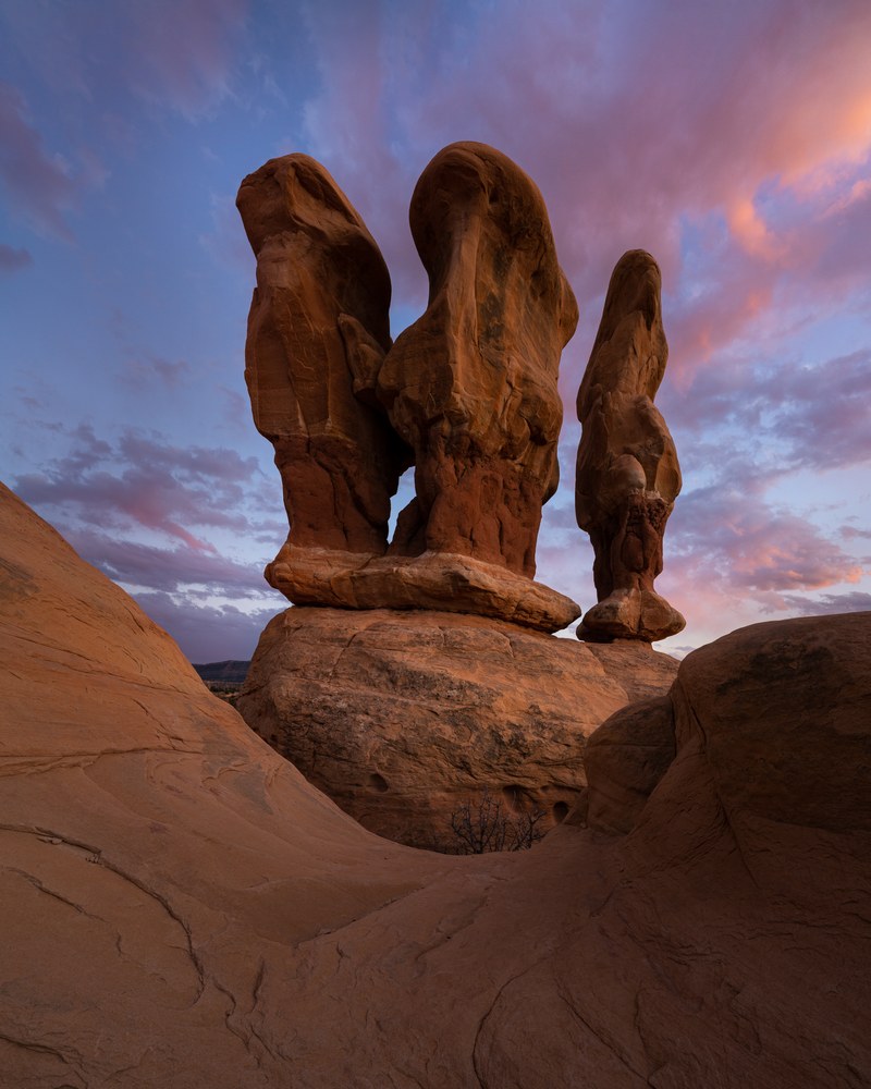 Three rounded hoodoos stand on a sandstone pedestal against pink and blue dusk cloud.