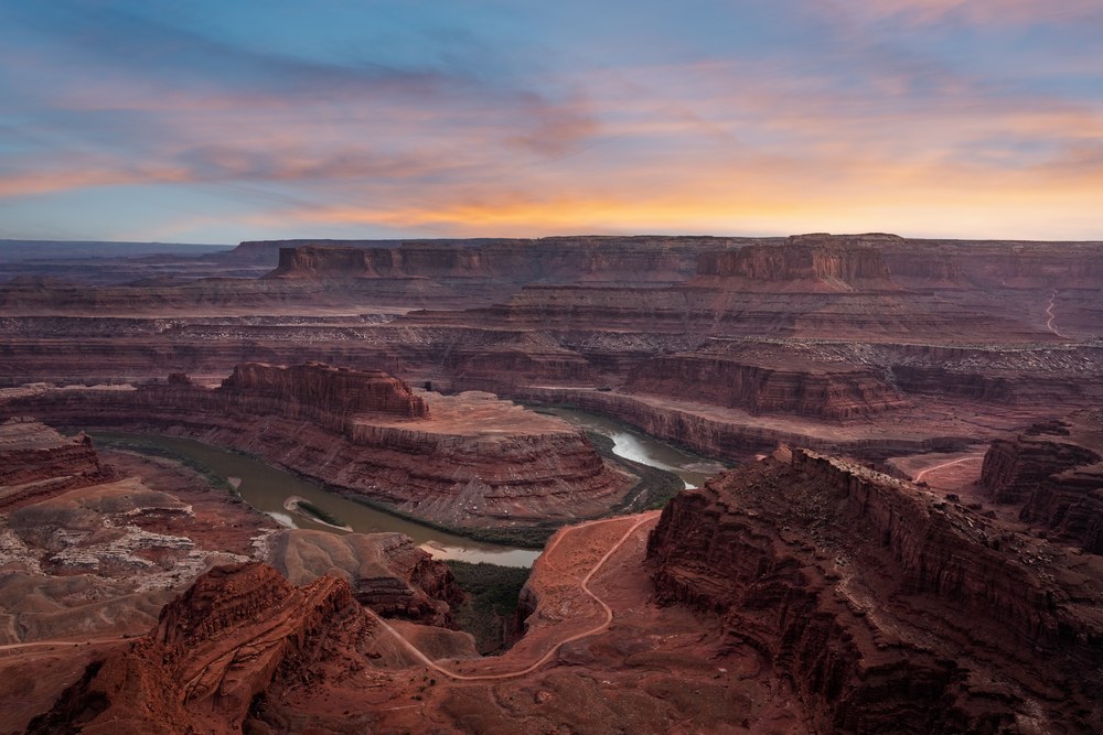 From a high rim, a green river bends through stacked red mesas and benches under a pale orange sunrise.