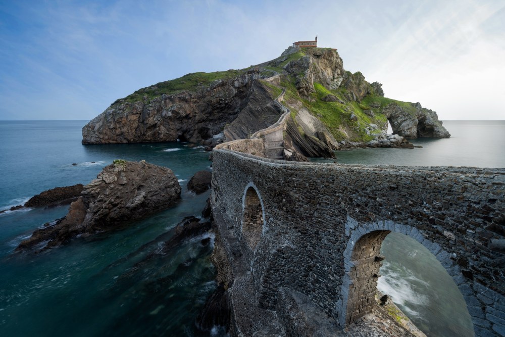 A stone causeway of arches climbs toward a green rock island crowned by a small chapel, calm blue sea on either side.