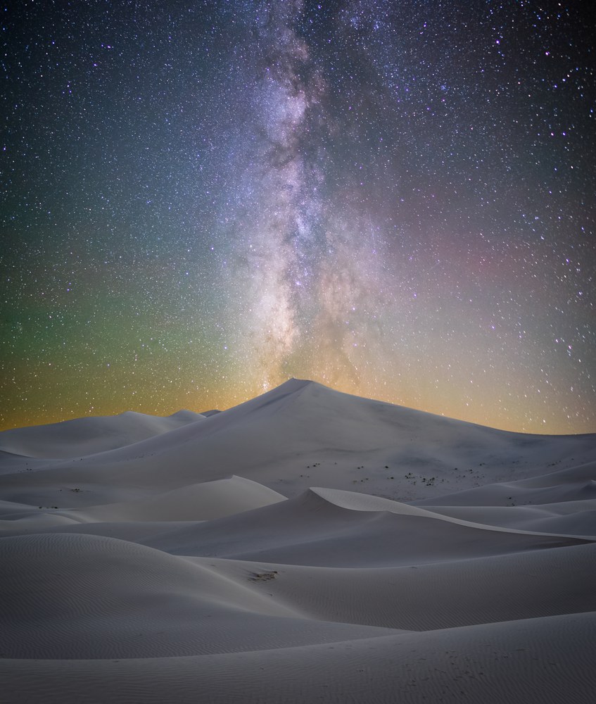 The Milky Way stands over pale dunes at night, faint green and gold airglow along the horizon behind them.