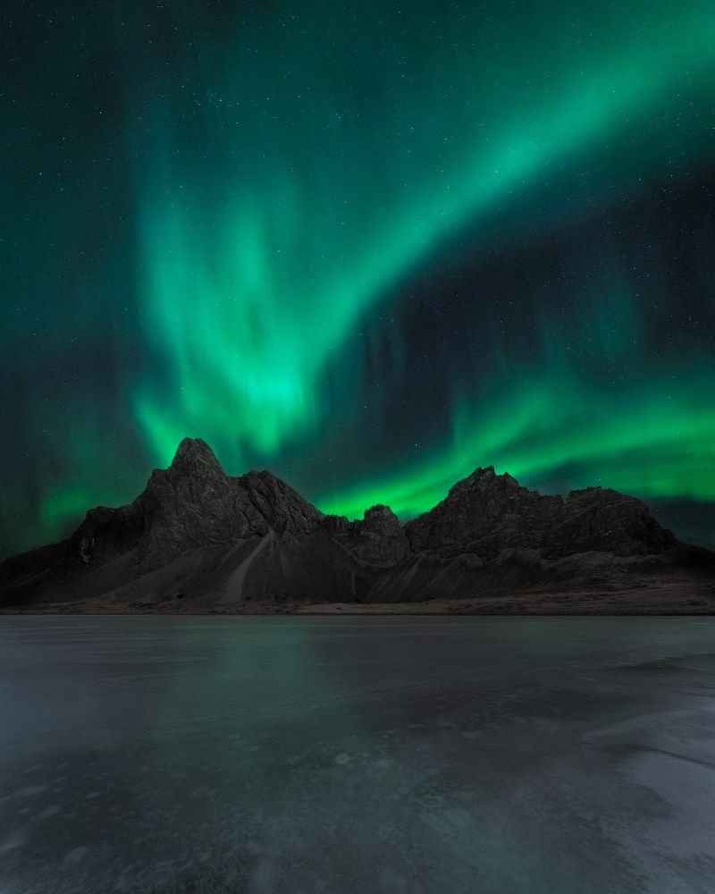 Green aurora pours down behind a range of jagged black peaks, mirrored faintly in a flat sheet of ice below.