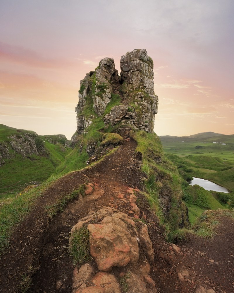 A worn earth path climbs a grassy ridge toward a rocky pinnacle, rolling green hills and a small loch beyond under a pastel sky.