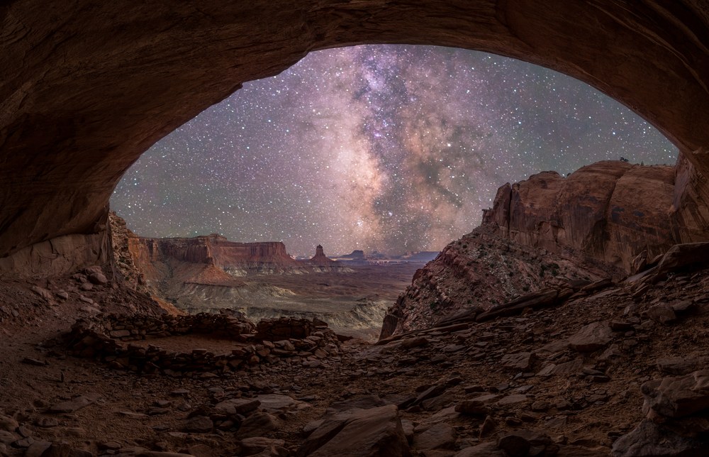 From inside a rock alcove, the Milky Way stands over distant canyon buttes, a low ring of stacked stones on the floor in front.