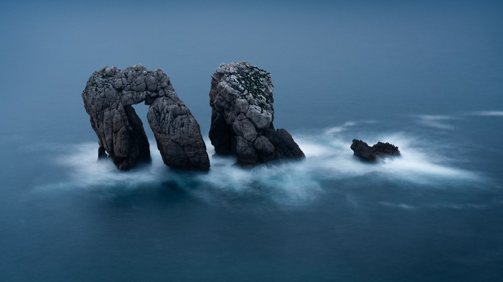 Two weathered rock pillars stand like a gateway in a flat blue sea, waves blurred to mist around their bases.