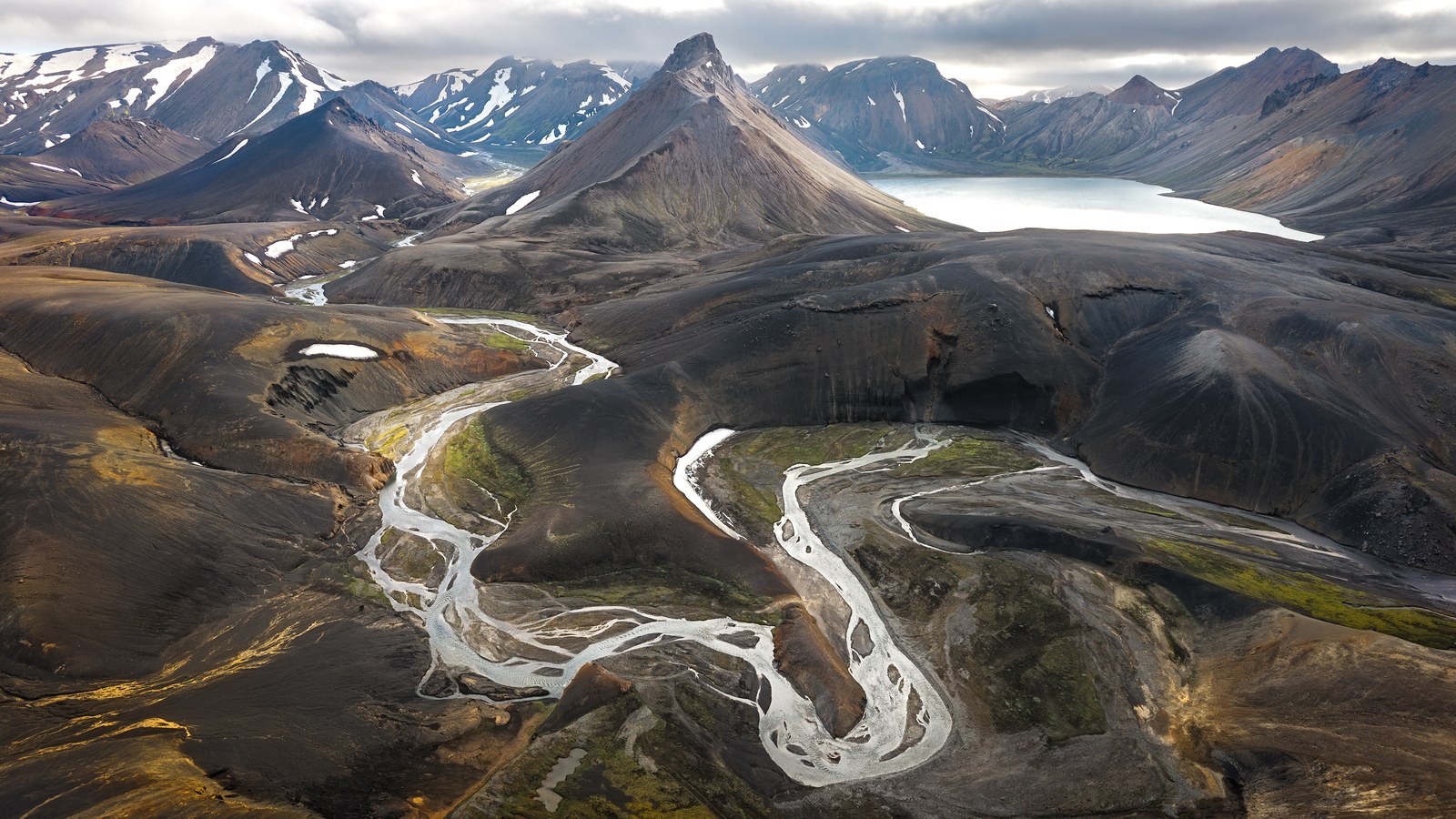 An aerial of a braided silver river winding through dark volcanic hills toward a pointed peak and a pale lake beyond.