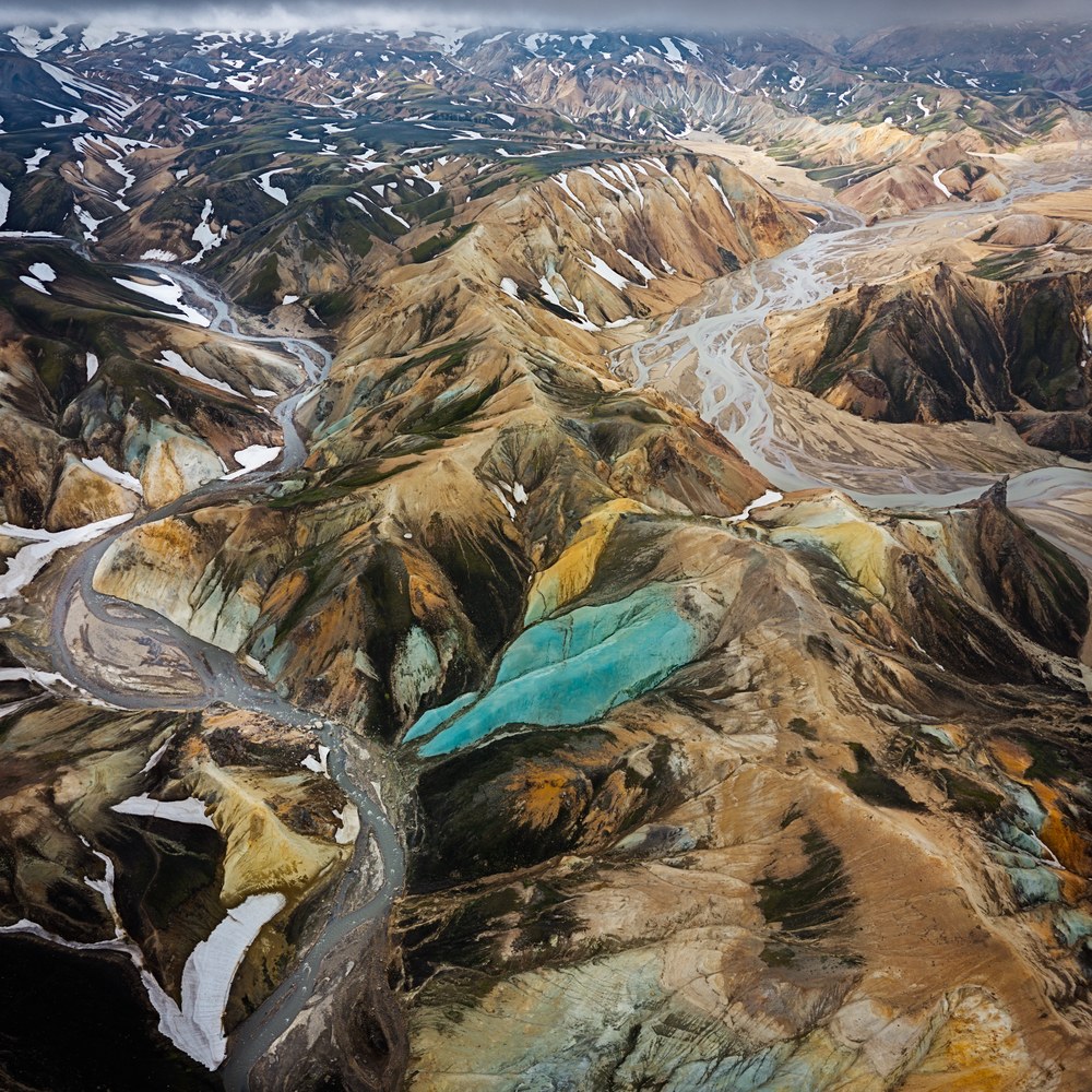 An aerial of banded rhyolite ridges in ochre, green and turquoise, braided grey rivers threading the valleys between them.