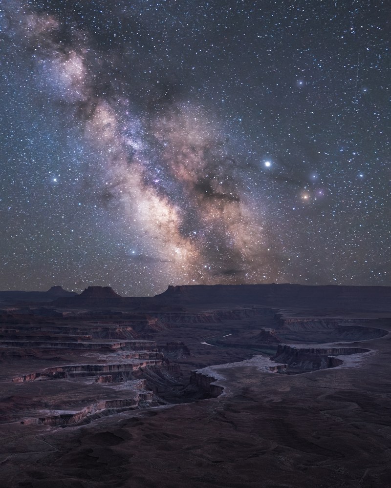 The Milky Way climbs above stepped canyon benches, a pale river winding through the flats far below.