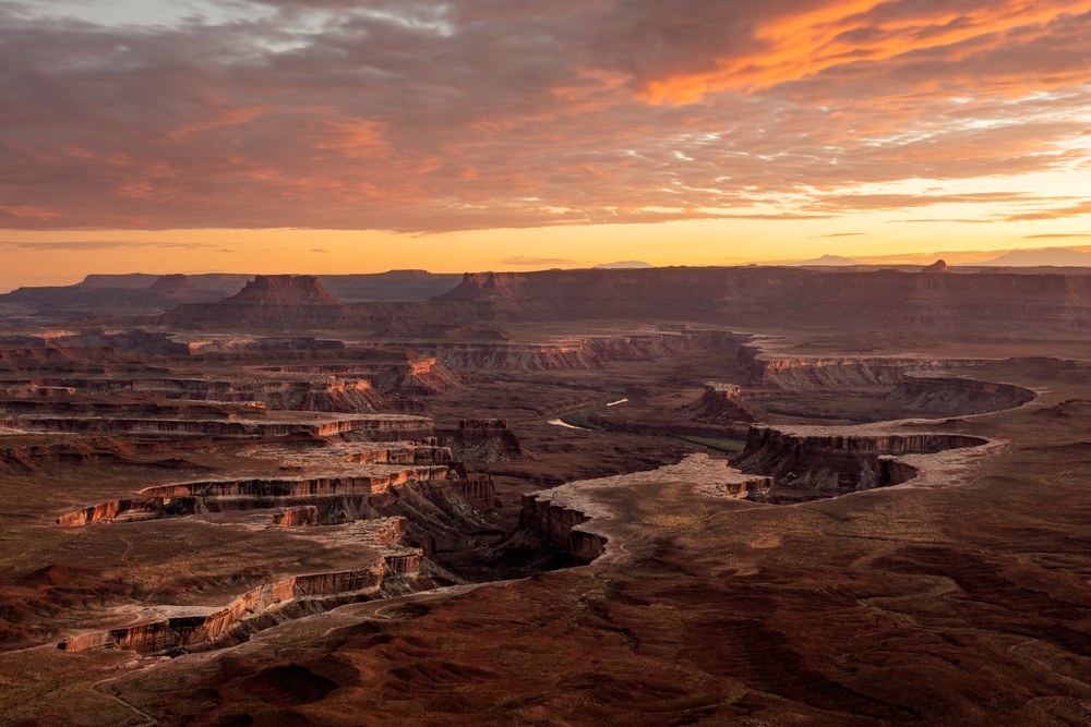 Orange fire runs through the cloud over a broad canyon, the river threading pale between stepped red benches below.