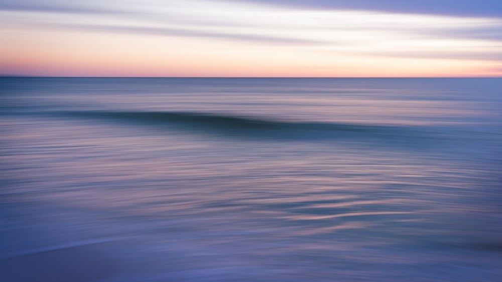 A single low wave reads as a soft horizontal band across water blurred by camera movement into stripes of pink and blue.
