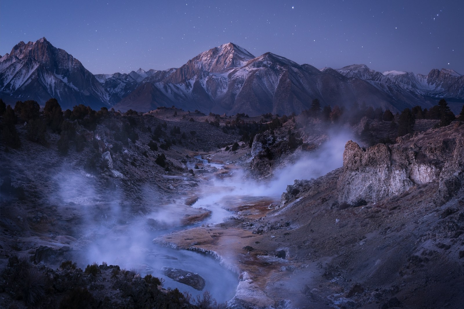 Steam rises off a geothermal creek winding through scrub toward snow-dusted peaks under a deep blue, star-flecked sky.