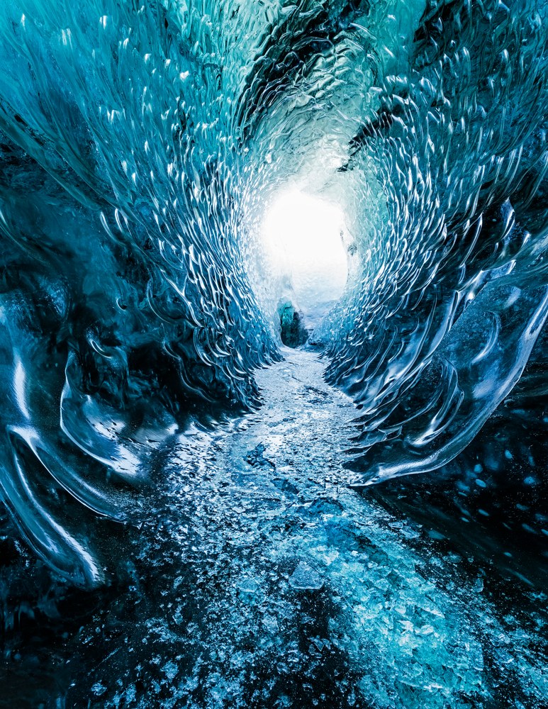Inside a glacier, a rippled blue ice tunnel narrows toward a bright opening, its floor covered in broken ice and grit.