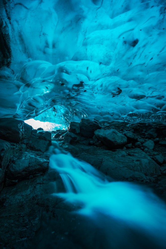 A rippled blue ice ceiling arches over dark rock as meltwater runs out toward a small bright opening.