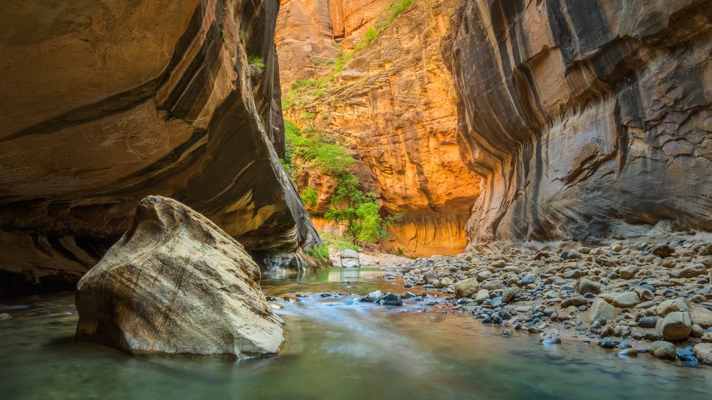 A large pale boulder sits in a shallow river inside a narrow canyon, the far wall glowing orange with reflected light.