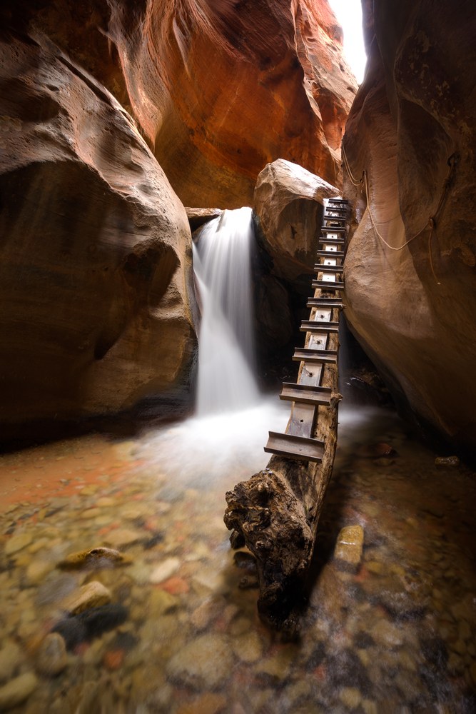 A wooden ladder is fixed to a fallen log beside a small waterfall in a narrow red slot canyon, coloured cobbles under the shallow water.
