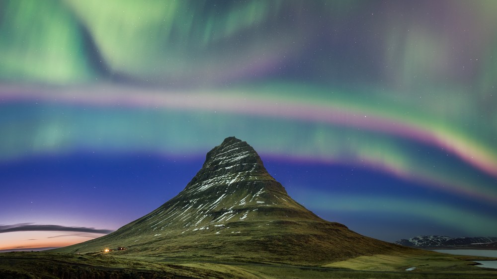 Bands of green and violet aurora sweep across the whole sky above a steep cone-shaped mountain.