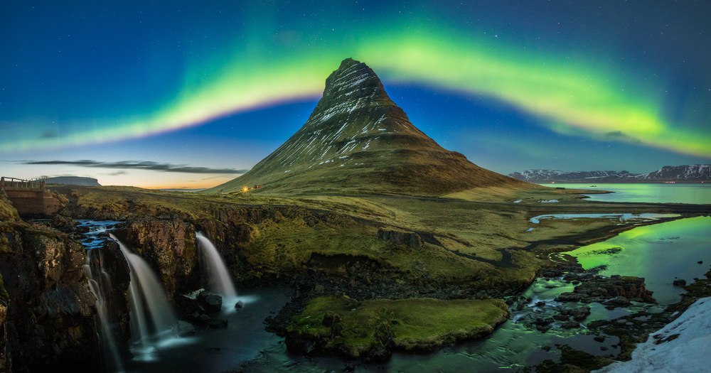 An arc of green aurora curves over a cone-shaped mountain while three small waterfalls drop over rock in the foreground.