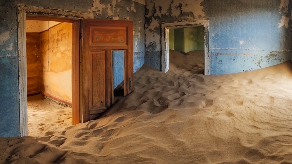 Sand drifts waist-deep through the doorways of an abandoned room with peeling blue and ochre walls.
