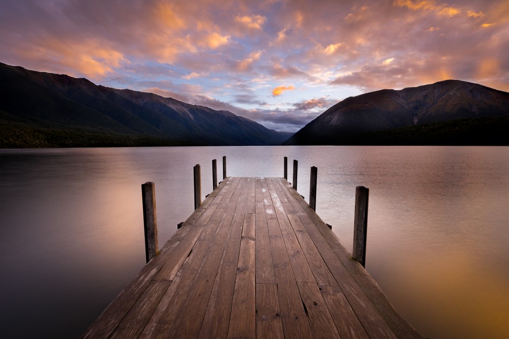 A wooden jetty runs straight out into a calm lake ringed by mountains, orange cloud overhead.