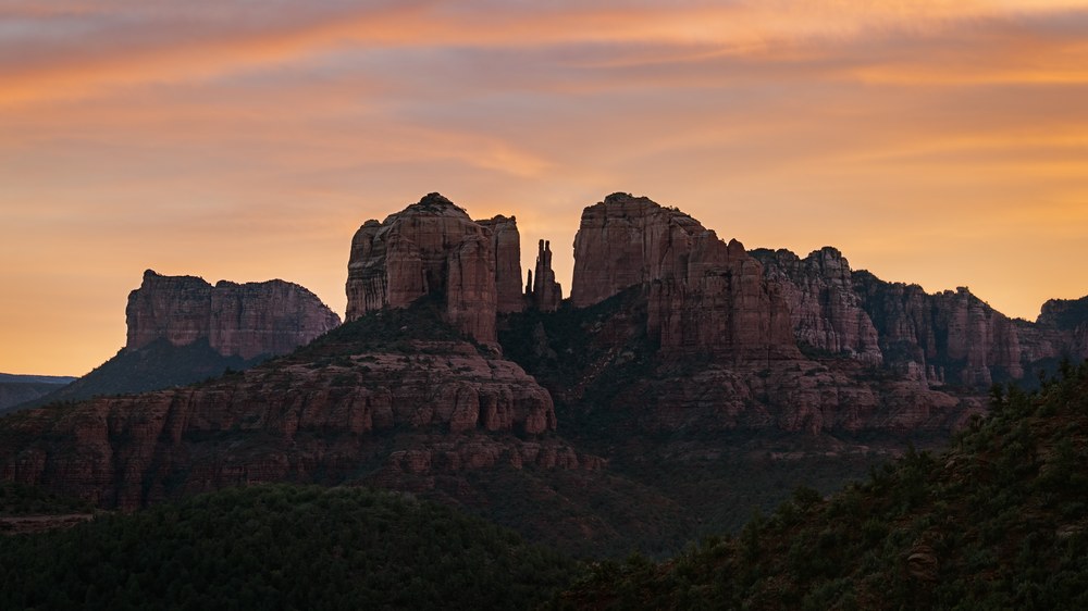 A broad red rock formation with a cluster of spires stands over dark treetops under a wash of orange sunset cloud.