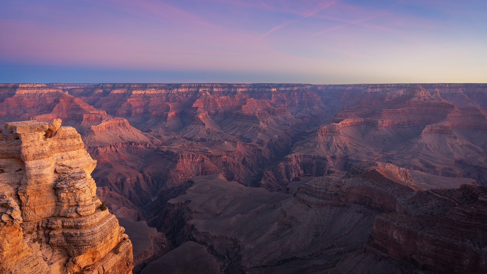 Ranks of layered canyon buttes recede to a flat horizon in cool dawn light, a rock outcrop at the left edge.