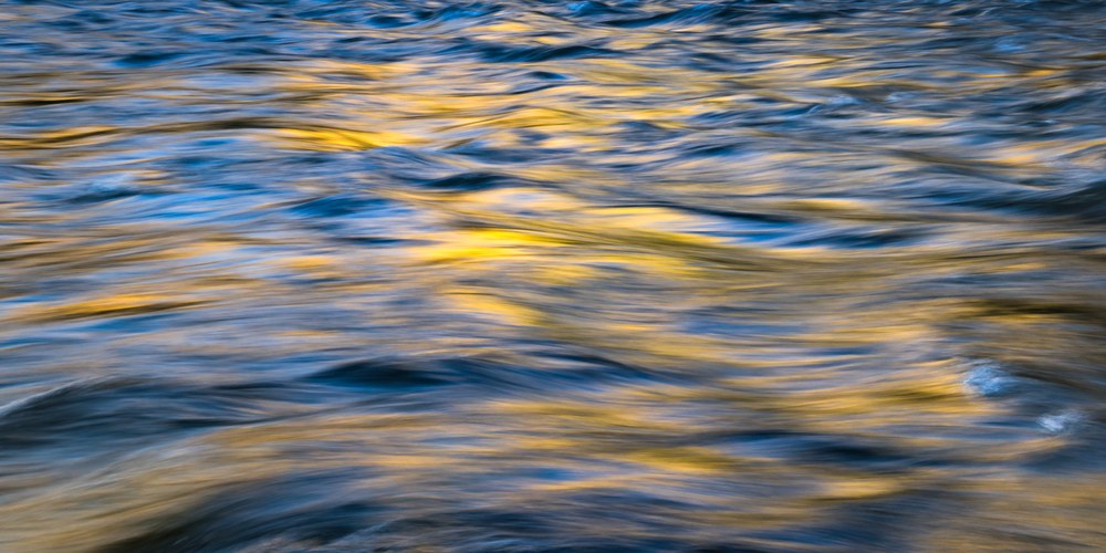 A close view of moving river water where gold and deep blue reflections smear into brushstroke shapes.