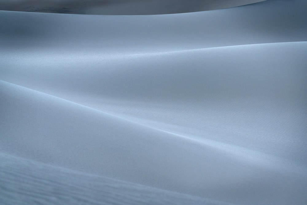 Soft pale curves of sand in flat blue-grey light, a single ridge line running diagonally across the frame.