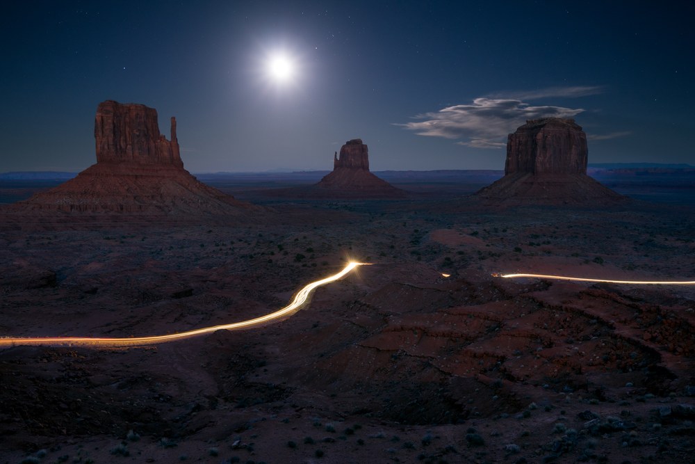 A full moon lights three desert buttes while a headlight trail draws a bright line across the scrub in front.