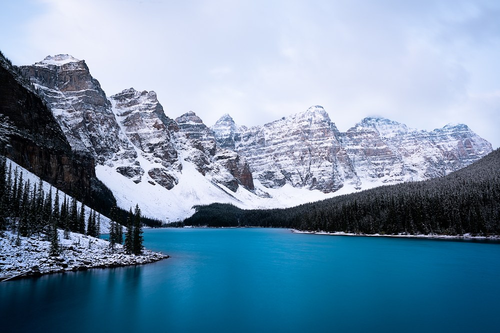 A wall of snow-dusted peaks rises above a turquoise lake edged with dark conifers after fresh snow.