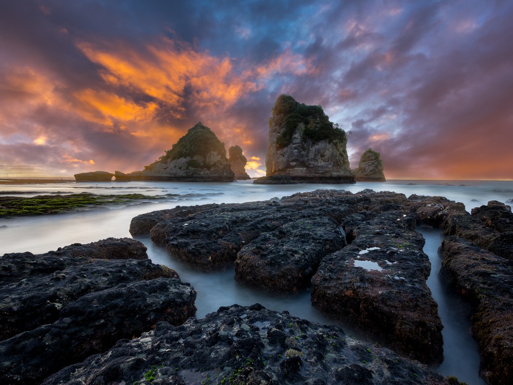 Dark blocky reef fissured with tide pools leads out to vegetation-topped sea stacks under orange and purple sunset cloud.