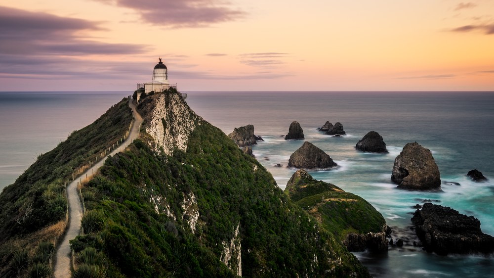 A white lighthouse caps a narrow green headland with a footpath climbing its spine, rock islets scattered through the sea beyond at sunset.