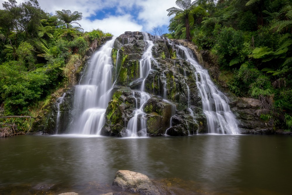 A waterfall splits into three sections down a mossy rock face into a still pool, tree ferns crowding both sides.