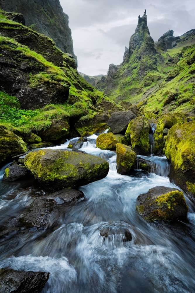 A stream runs fast over moss-covered boulders through a bright green gorge, a jagged pinnacle standing at its head.