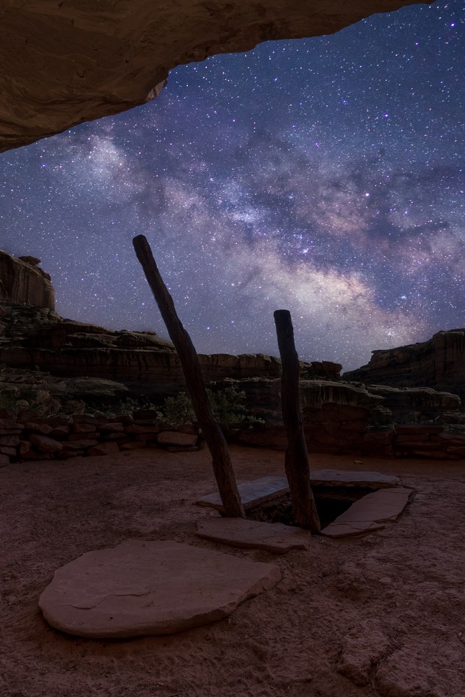 Two wooden poles rise from a hatch in a kiva roof beneath a rock overhang, the Milky Way filling the canyon beyond.