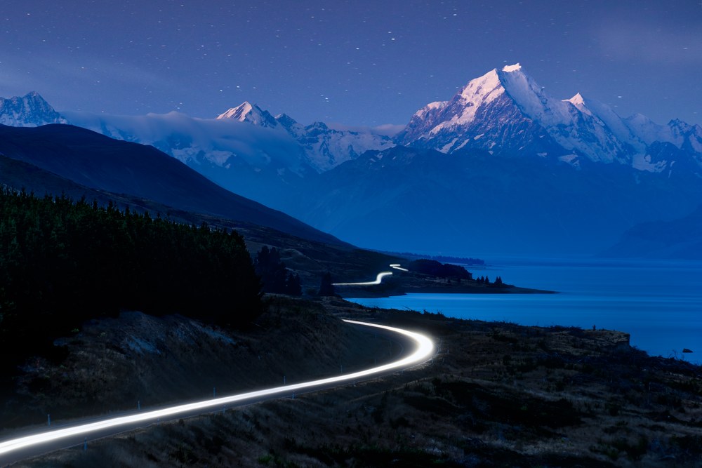 A headlight trail draws a bright curve along a road above a blue lake, a snow-capped peak catching the last light under stars.