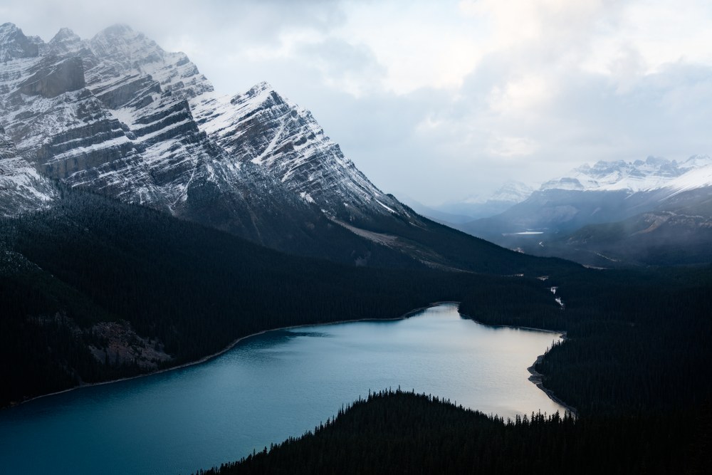 A teal lake lies between dark forested slopes below a snow-streaked peak, the valley receding into cloud.